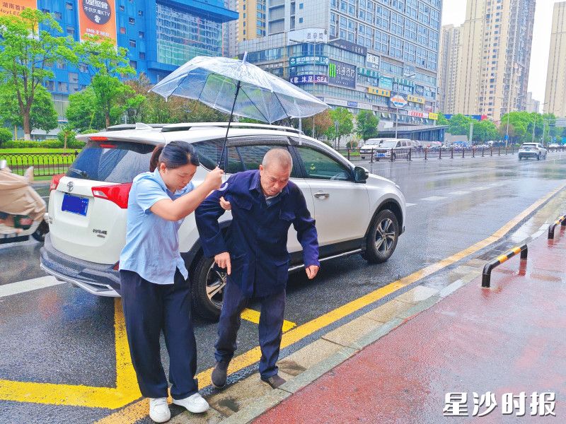 工作人员撑着雨伞，小心翼翼搀扶着老人到公交站台。